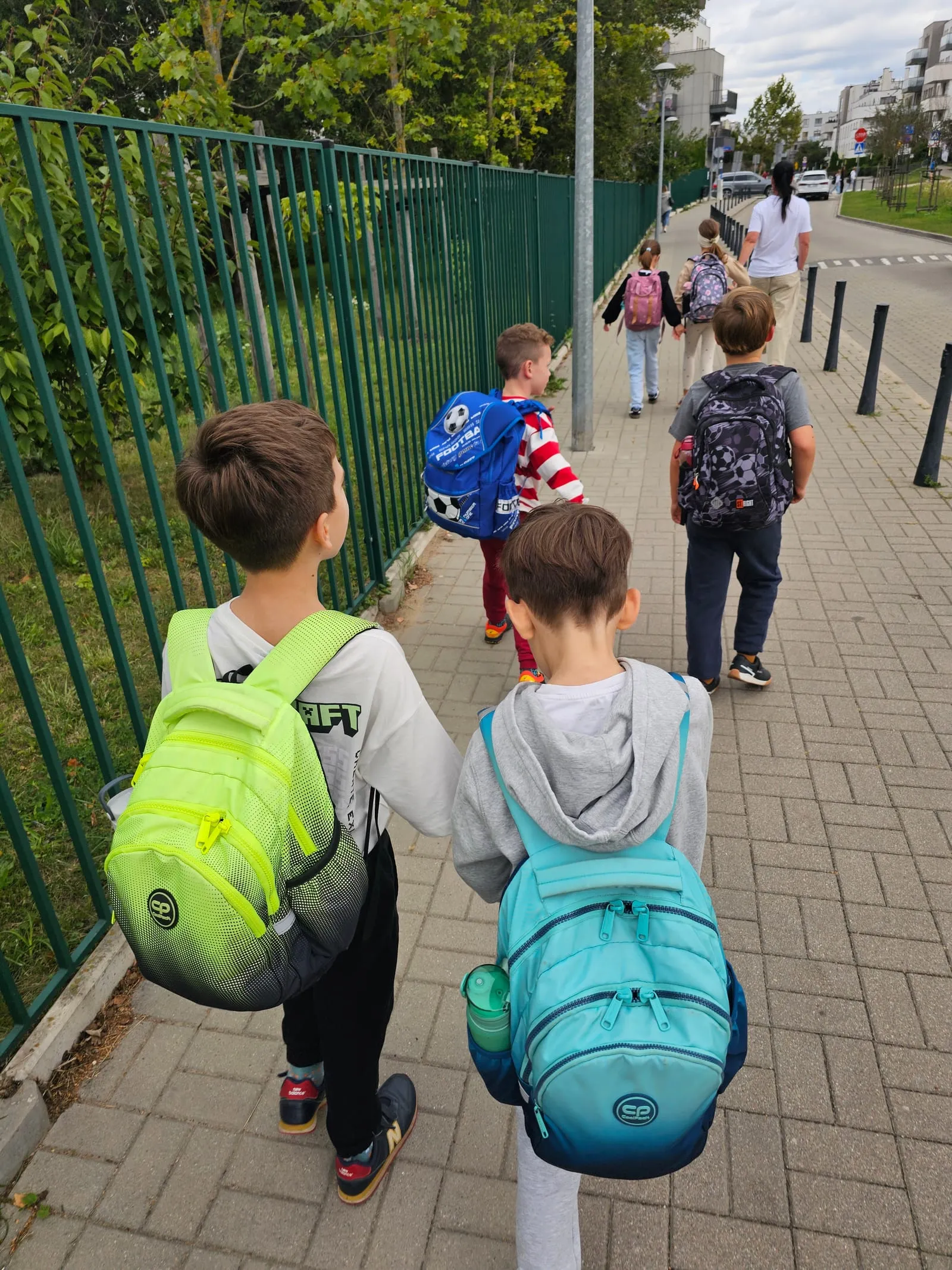 Dzieci z plecakami idą chodnikiem na zajęcia, widziane od tyłu